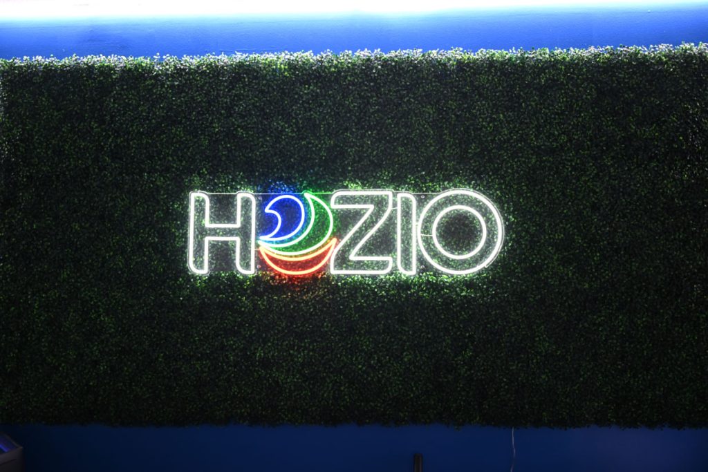 Hozio neon sign on green artificial hedge wall, vibrant lighting for modern branding display.