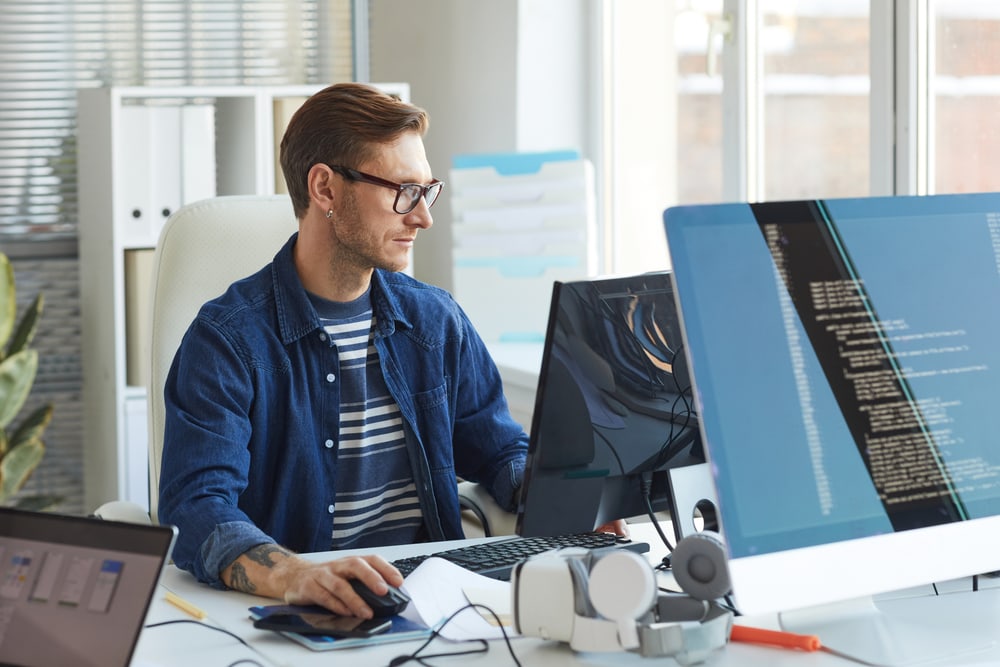 Focused male developer working on computer coding at modern office desk with multiple screens.